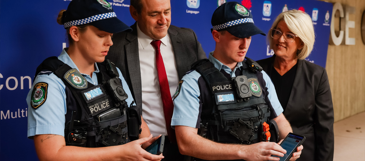 Photo of 2 police officers both using a mobile device in front of officials in a conference