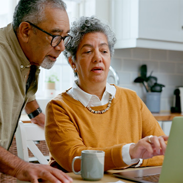 Photo of couple in a kitchen, one seated at a table using a laptop with a mug in front, the other standing beside and looking at the screen