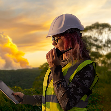 Photo of a person in hard hat and reflective vest using a tablet and field radio with sunset and trees in background