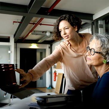 Photo of two people in an office, one pointing at a laptop screen while the other looks on