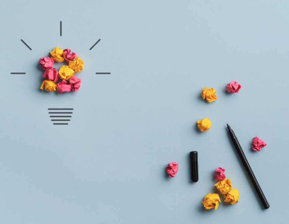 Photo of a creative concept showing a light bulb formed from crumpled paper balls on a blue background, with black lines illustrating rays of light. A pen and additional paper balls are scattered to the right