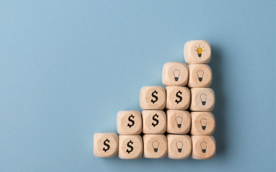 Photo of wooden blocks with dollar signs and lightbulbs arranged as stairs, symbolising investment leading to innovation
