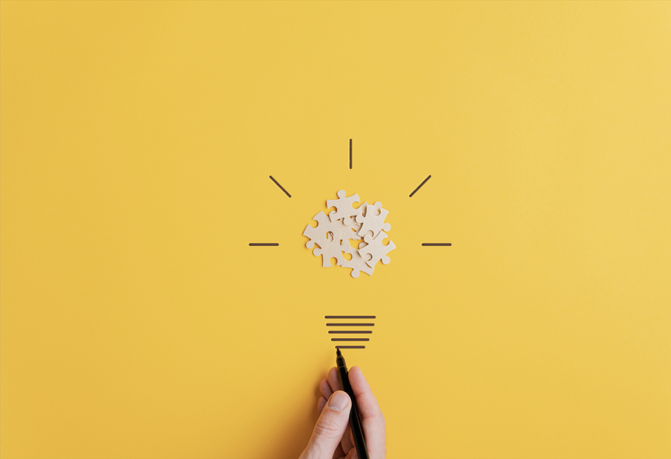 Photo of a hand drawing below some puzzle pieces forming a light bulb on a yellow background
