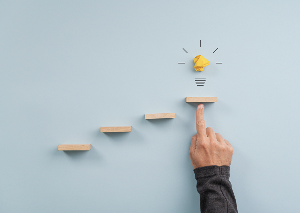 Photo of a hand pointing to top step of wooden block staircase with a crumpled paper light bulb above, symbolising reaching an idea
