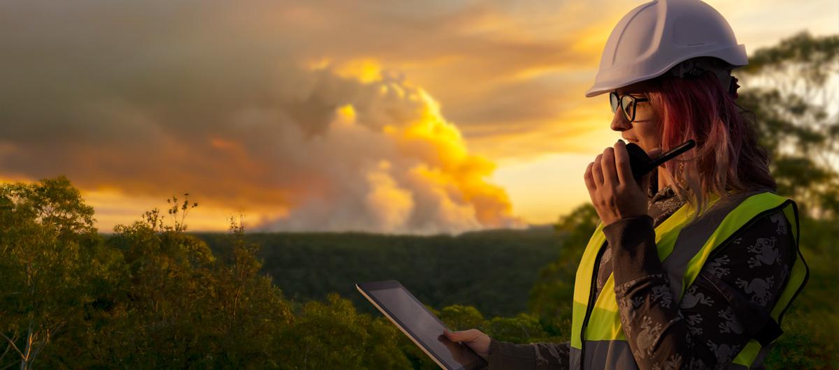 photo of a person in safety gear using a tablet and radio near a smoky forest at sunset.
