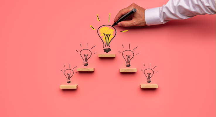 Photo of a hand drawing a large light bulb above wooden blocks arranged in a pyramid, with smaller light bulbs on lower steps, symbolising idea development