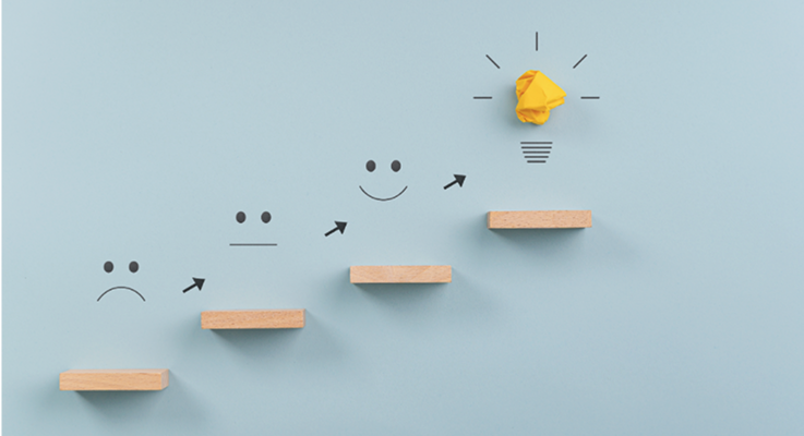 Photo of wooden staircase with arrows progressing from sad to happy faces, ending in a crumpled paper light bulb on a blue background