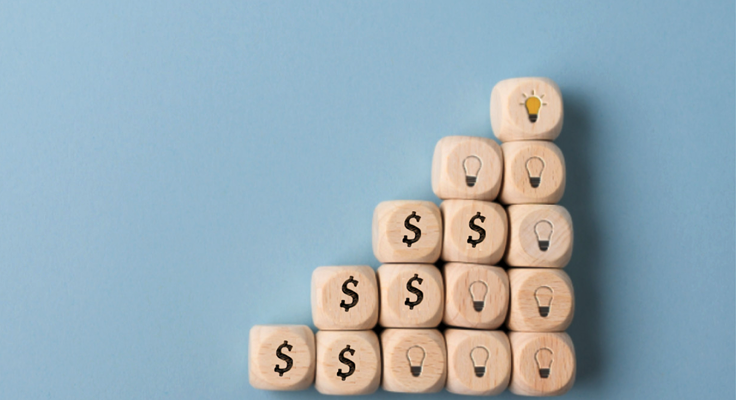 Photo of wooden blocks with dollar signs and lightbulbs arranged as a pyramid, ending with a lit bulb to symbolise financial growth and innovation